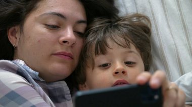 Yatakta Nişanlı Çocuk ve Anne, Çevrimiçi İçeriği Gösteren Çocuk Olarak Telefon Ekranına Odaklanmış