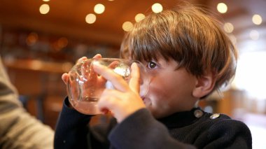Küçük çocuk restoranda su yudumluyor. Çocuk içki içiyor ve elinde cam tutarak su içiyor.