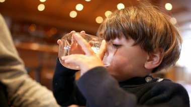 Küçük çocuk restoranda su yudumluyor. Çocuk içki içiyor ve elinde cam tutarak su içiyor.