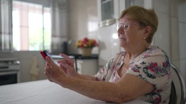 Üst düzey bayan sosyal medyada ev mutfağındaki telefonu kullanıyor. Beyaz kadın akıllı telefon cihazı tutuyor.