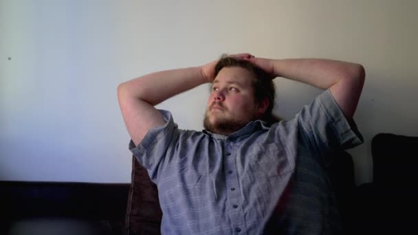 一个沉思的 胖乎乎的男人在屋里思考 双手放在脑后沉思着 坐在沙发上的心事重重的人 — 图库视频影像
