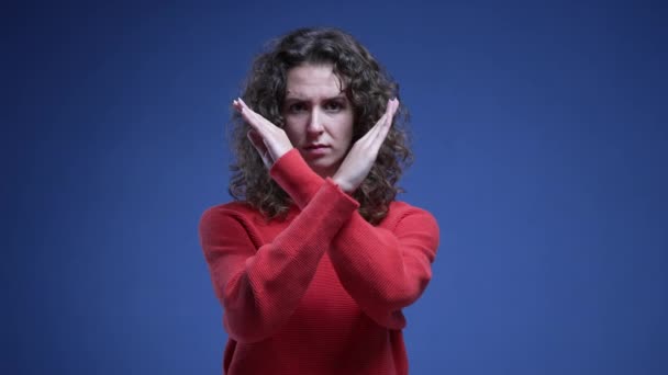 若い女性は指を指さしてカメラにノーと言います 青い背景に立っているオファーや行動を拒否する腕で十字架を作る20代の女性 — ストック動画