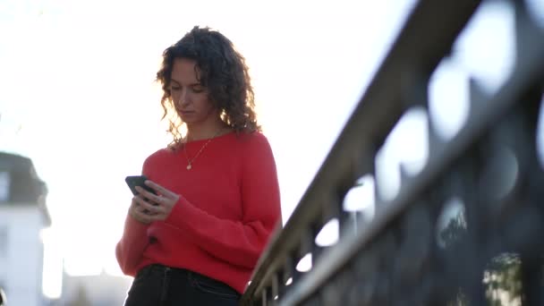 市街地の外で 金属製の鉄道を日光で走らせている 携帯機器を見ている女性です 携帯電話でテキストメッセージを送信する20代女性 — ストック動画