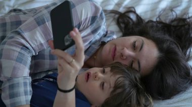 Rahat anne ve oğul pijama giyerek, yatakta telefonla meşgul olarak, çocuk ve anne arasında geçen ve internetteki eğlence içeriğine bakan