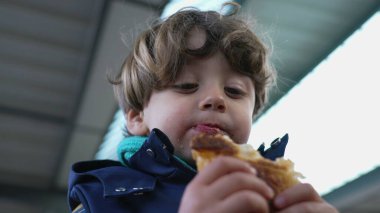 Tren istasyonunda kruvasan yiyen neşeli çocuk, sonbahar ceketi ve atkısı giymiş.