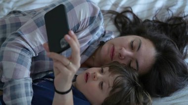Rahat anne ve oğul pijama giyerek, yatakta telefonla meşgul olarak, çocuk ve anne arasında geçen ve internetteki eğlence içeriğine bakan