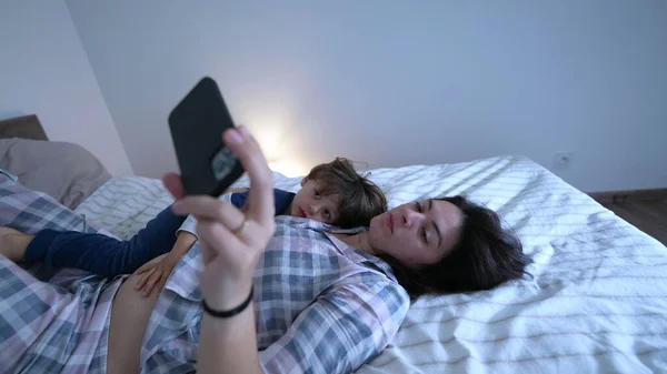 Mother and child laid in bed staring at cellphone device screen. Parent holding phone with one arm watching content online with son together wearing pajamas