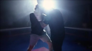 Dövüşçüler, boks ringinin içinde, dramatik ışıklandırmalarla diz yumruklaşıyorlar. Muay Thai dövüşçüleri rakiplerinin yumruk ve tekmelerini önlemek için birbirlerini sıkıca kavrıyorlar