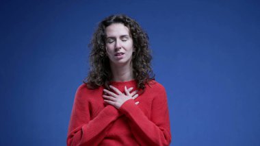 Genç bir kadın elini göğsüne koyarak rahatlıyor. Mavi arka planda durarak mücadele ettikten sonra derin bir nefes alıyor.