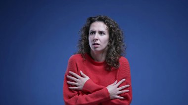 Korkmuş genç bir kadın korkusunu göstermekte tereddüt ediyor ve kırmızı süveter giyerek mavi arka planda panikliyor. 20 'li yaşlarda kadın.