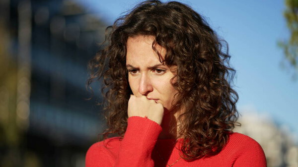 Занятая молодая женщина с подбородком, стоящая в городе во время заката в глубоком созерцательном самоосмотре, чувствуя страх и тревогу.