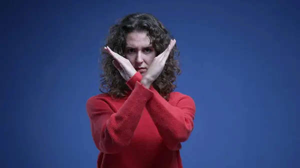 Young woman saying NO to camera by pointing and waving finger. 20s female person making cross with arms rejecting offer or behavior standing on blue backdrop