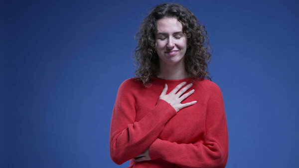 Grateful woman putting hands on chest feeling gratitude and faith on blue backdrop