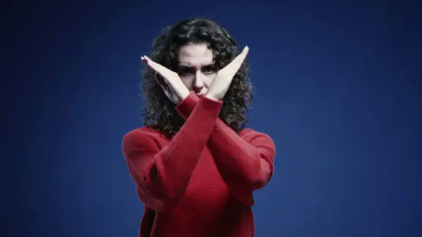 Young Woman Firmly Saying NO to Camera, 20s Woman Rejecting with Crossed Arms and Finger Wave, standing on blue backdrop