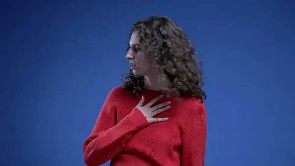 Young woman feeling relief by putting hand on chest, taking a deep breath of relaxation after struggle standing on blue background
