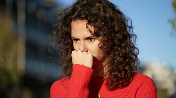 Занятая молодая женщина с подбородком, стоящая в городе во время заката в глубоком созерцательном самоосмотре, чувствуя страх и тревогу.