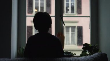 Kış mevsiminde ev penceresinden düşen karlara bakan çocuk silueti. Aralık ayında, kasvetli bir günde, doğası gereği hipnotize edilen genç bir çocuğun arkası.