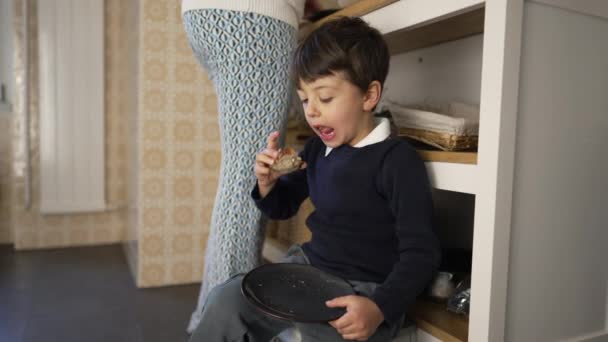 Small boy snacking bread with cheese seated on kitchen floor and leaning on furniture while mother stands in background cooking