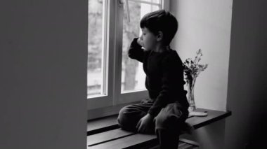 Sıkıcı küçük çocuk pencerenin kenarında oturmuş tek renkli gözleriyle üzüntüsünü ve çocukluk depresyonunu anlatıyor, siyah ve beyaz.