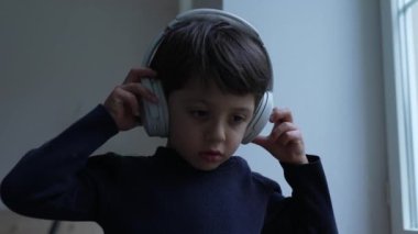 Kulağında kulaklıkla müzik, şarkı ya da ses dinleyen küçük bir çocuk evde pencerenin yanında duruyor. Çocuk gürültü önleyici kulaklık takıyor