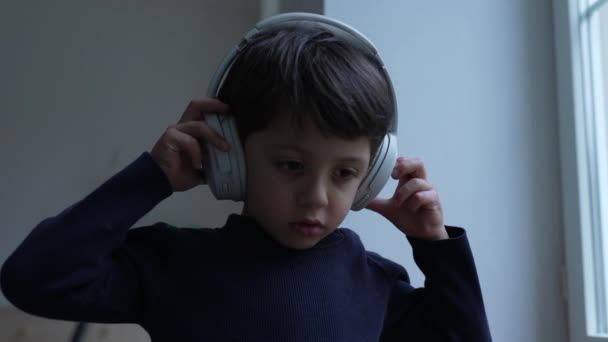 オーディオを聞く小さな少年が 家で窓に立って耳の上にヘッドフォンを持っています 子供はヘッドセットをキャンセルするノイズを身に着けています — ストック動画