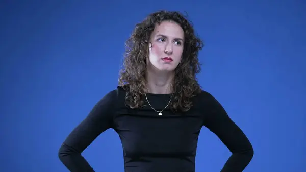 Annoyed woman rolling eyes in disgust and contempt while standing on blue background with hands on hips