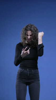 Cep telefonu cihazını söken kadın, mavi arka planda duran telefon ekranında harika haberleri kutluyor.