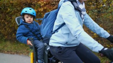 Çocuk ve anne sonbahar sezonunda birlikte bisiklete biniyorlar. Bisikletli çocuk yeşil yolda arka koltukta