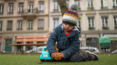 Sonbaharda Park 'ta kamyonet kıskacıyla oynayan, soğuk sezonda bereli ceket giyen ve oyuna dalmış bir çocuk.