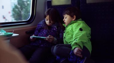 Passenger Small girl playing game with handheld console while younger brother stares at her screen play, kids engaged with technology while on the go inside train