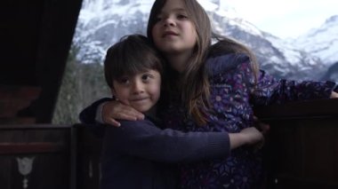 Küçük kardeş dağ evinde kız kardeşini kucaklıyor. Kış kayak sezonu boyunca arka planda karla kaplı İsviçre dağlarıyla birlikte.