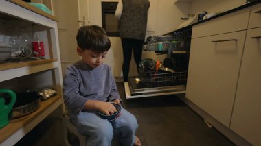 Genç bir çocuk mutfakta oturmuş yaban mersini yerken anne mutfak gerisindeki bulaşık makinesine mutfak gereçleri ve tabakları yerleştiriyor. Günlük aile sahneleri.