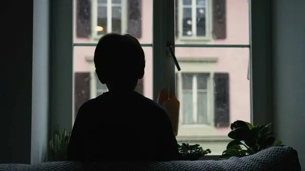 Silhouette of child staring at snow fall from home window during winter season. Back of young boy hypnotized by nature during moody day, December season