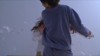 Çocuklar yastık savaşının ortasında yataklarında tüyleri uçuşan ve hız rampasında 1000 fp hızla çekilen bir kamerayla yakalanan çocuklar.