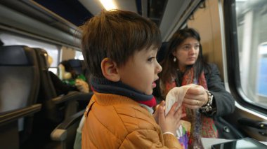 Küçük çocuk annesiyle trenle seyahat ediyor. Atkı ve ceket giyen çocuk manzaraya bakıyor. Yüksek hızlı ulaşım aracının içinden geçiyor.