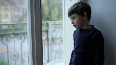 İçgözlemci çocuk, pencerenin kenarında durmuş üzgün ve düşünceli bir ifadeyle apartmandan dışarı bakıyor.
