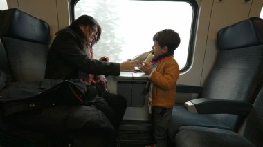 Anne ve çocuk trenle seyahat ediyor, küçük çocuk muz yiyor, anne oğluna içecek su veriyor, aile tatillerinde seyahat ediyor.