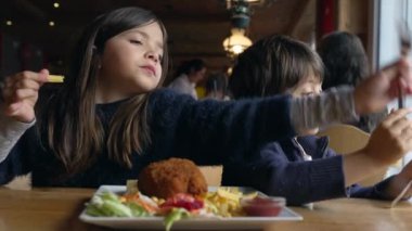Lokantada yemek yiyen çocuklar, kardeşler. Küçük erkek ve kız kardeşler, ahşap iç mekandaki lokantada patates kızartmasının ve tavuk yemeğinin tadını çıkarıyorlar.