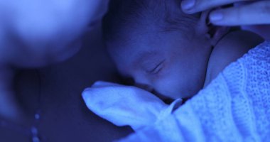 Mother holding newborn baby at hospital next to violet phototherapy lamp, real life affection and love