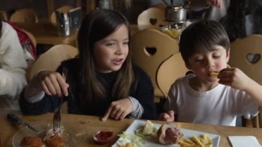 Çocuklar lokantada yemek yiyor. Küçük kardeşler, lokantada kız ve erkek kardeşler yemek yiyor. Bir tabak kızarmış Milano eti.