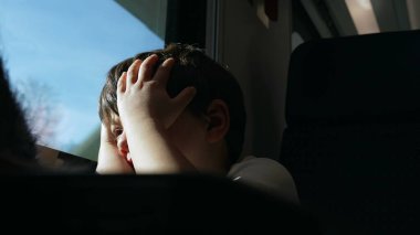 Exhausted child rubs eyes and face while on a moving train, passenger kid wakes up from nap while traveling feeling tired by window