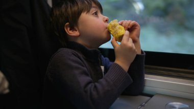 Thoughtful little boy snacking corn while looking at scenery pass by from inside train