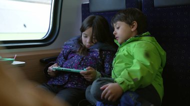 Little girl playing with video game while younger brother stares at screen while traveling by train. Children engaged with virtual play while on commute inside high speed transportation