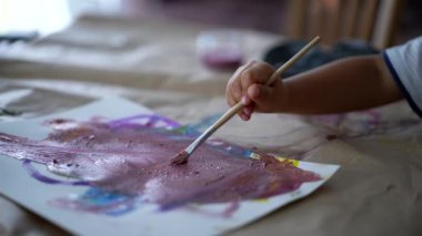 Boyalı fırçayı tutan, kağıt üzerinde suluboya soyutlama yapan yakın çocuk, evde sanatsal işlerle uğraşan 3 yaşında bir çocuk.