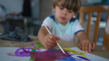 Sanatçı çocuk evde suluboya boyayla oynarken anne arka planda angarya işler yapıyor. Fırçayla resim yapan yaratıcı küçük çocuk.