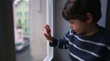 Pencereden dışarı bakan küçük bir çocuk. Çocuk ikinci kattaki apartman dairesinden sokağa bakıyor. Süveter giyen çocuk