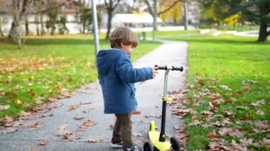 Küçük bir çocuk, sonbahar günü parkta dikilip mavi ceketini giyiyor ve 3 tekerlekli scooterını kontrol ediyor.