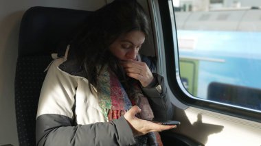Trenin içindeki cep telefonu cihazına bakan banliyö kadını pencerenin önünde oturuyor ve arka plan manzaralı. Akıllı telefon aygıtını kontrol eden yolcu