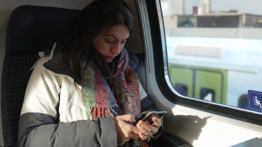 Trenin içindeki cep telefonu cihazına bakan banliyö kadını pencerenin önünde oturuyor ve arka plan manzaralı. Akıllı telefon aygıtını kontrol eden yolcu