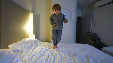 Mutlu genç 3 yaşındaki çocuk otel yatağında zıplıyor, kaygısız heyecanlı çocuk çarşaflarda zıplayarak gezerken eğleniyor.